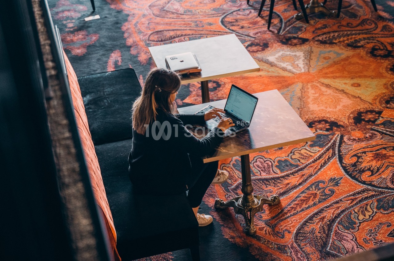 Une femme travaille sur un ordinateur portable, assise à une table dans un espace avec un tapis coloré.