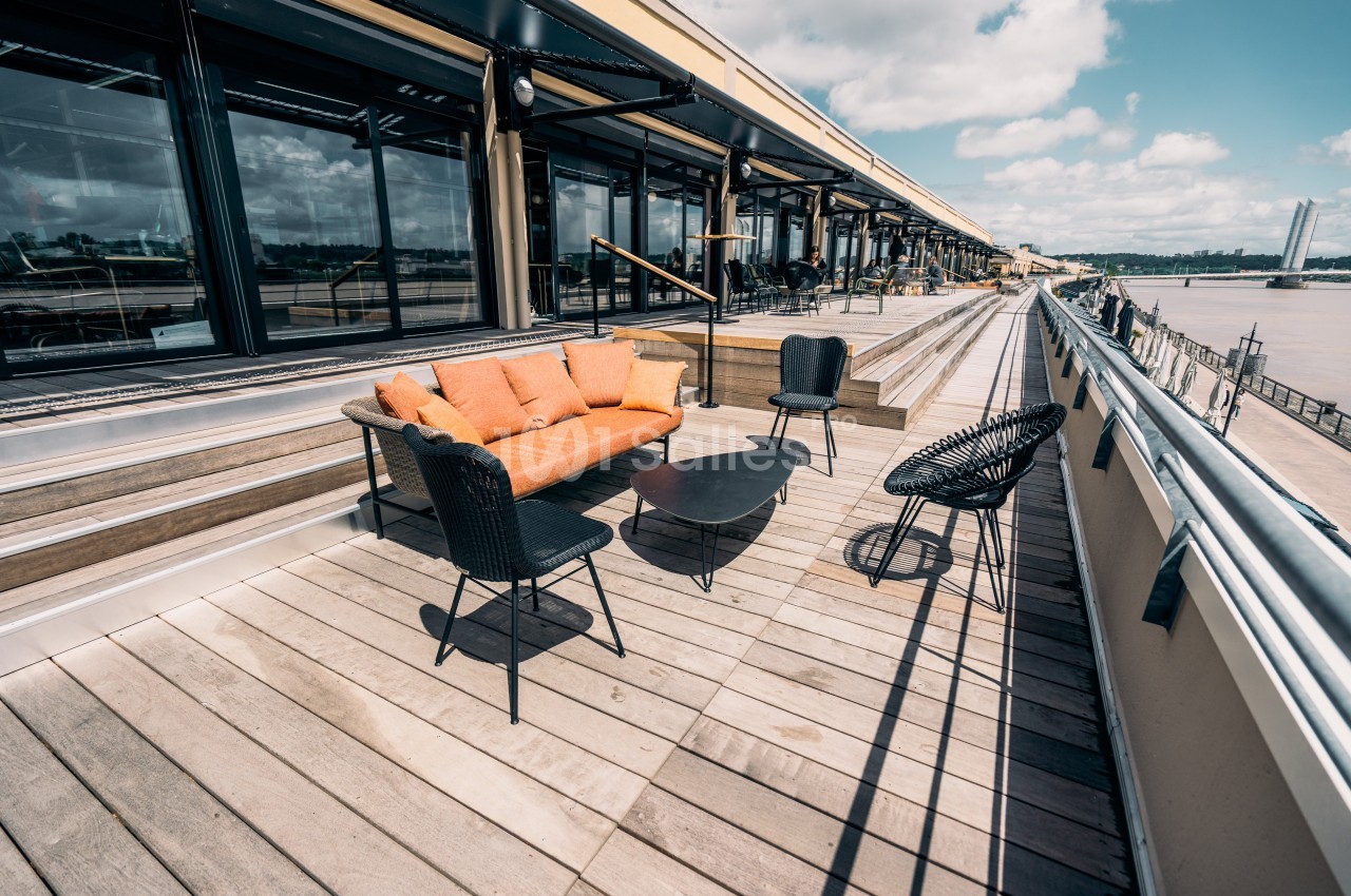 Terrasse en bois avec canapé orange, fauteuils noirs et table basse, donnant sur un espace extérieur ensoleillé.