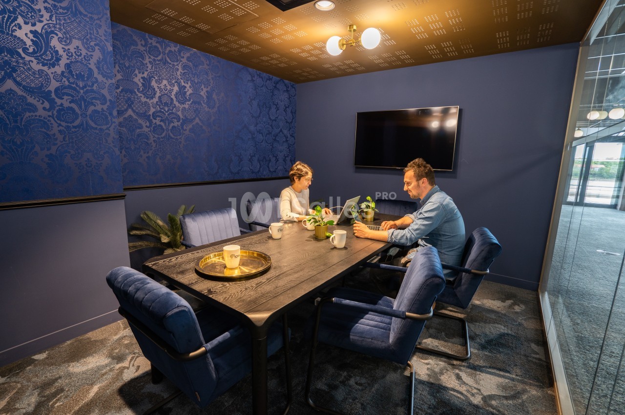 Deux personnes travaillent à une table dans une salle de réunion moderne avec murs bleus et éclairage chaleureux.