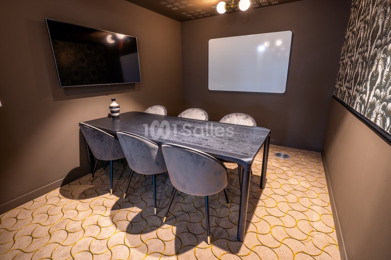 Salle de réunion avec table en bois foncé, six chaises grises, tableau blanc et écran mural.