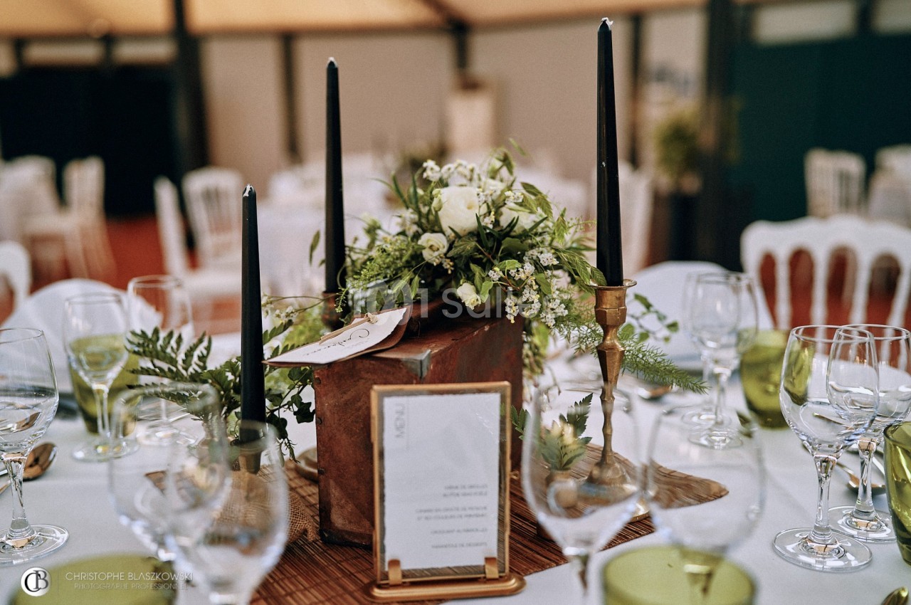 Centre de table avec fleurs blanches, bougies noires et éléments végétaux, entouré de verres et assiettes.