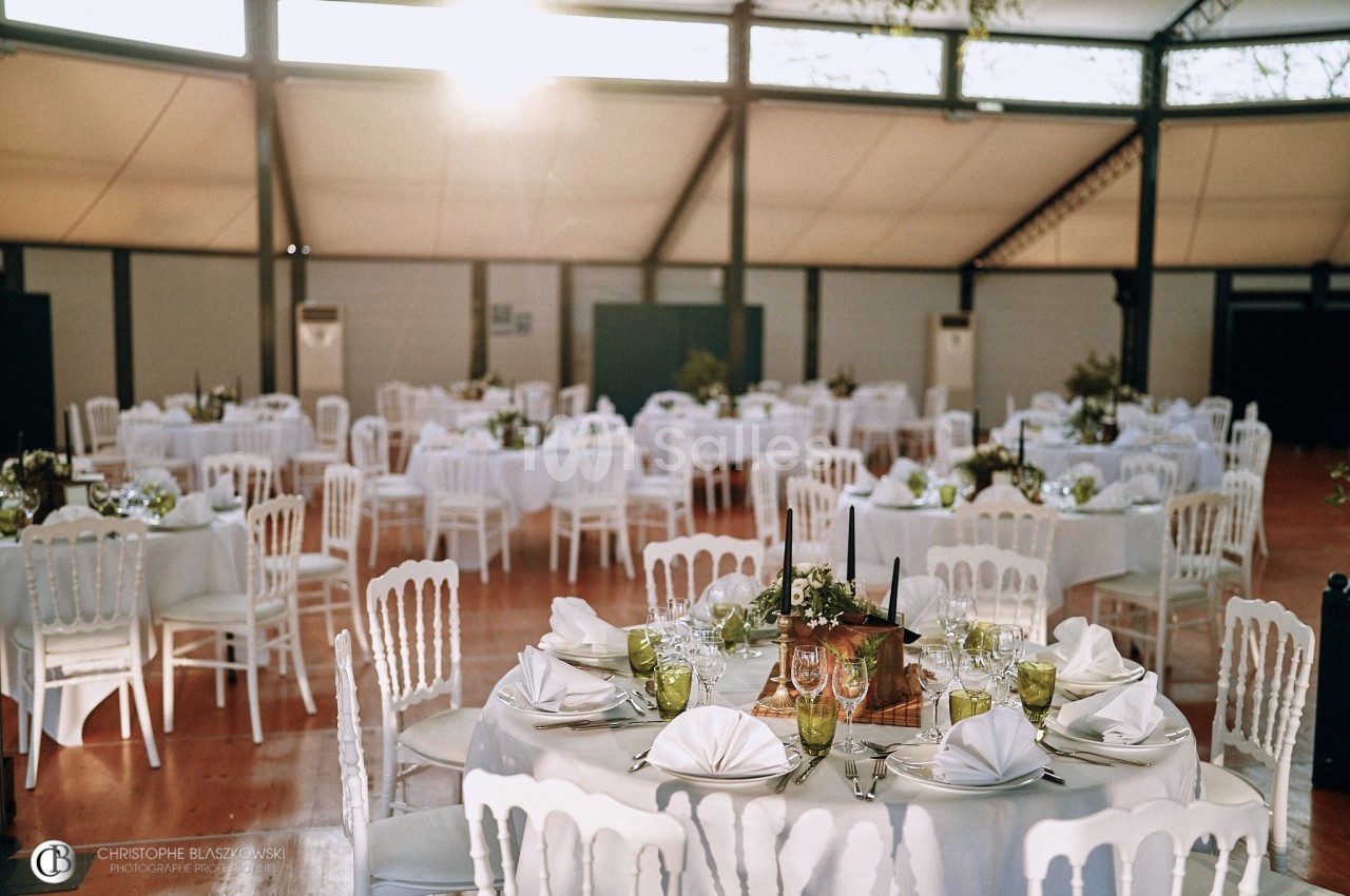 Salle de réception lumineuse avec tables rondes dressées, nappes blanches, chaises assorties et décorations florales.