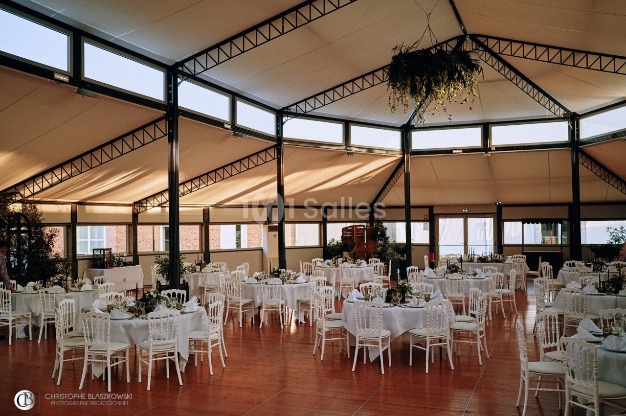Salle de réception lumineuse avec tables rondes dressées, chaises blanches et décoration végétale suspendue.