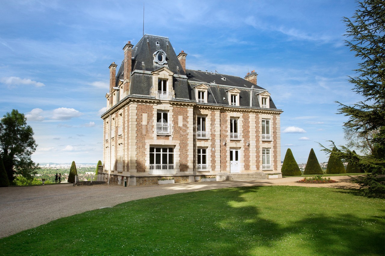 Façade d'un manoir en briques et pierre entouré d'un jardin avec pelouse et arbres, sous un ciel dégagé.