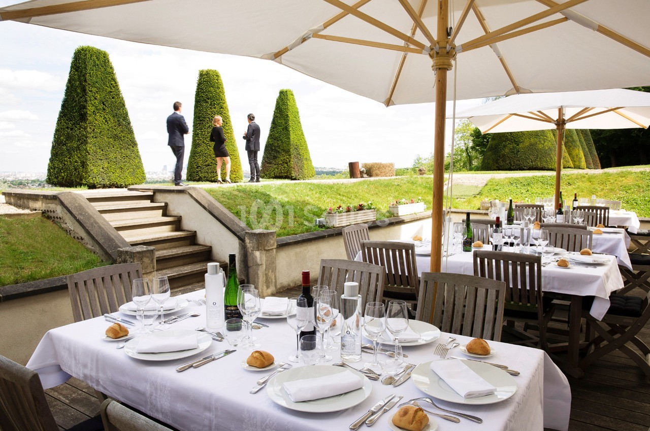 Terrasse d'un restaurant avec tables dressées, vue sur un jardin et trois personnes debout près de haies taillées.