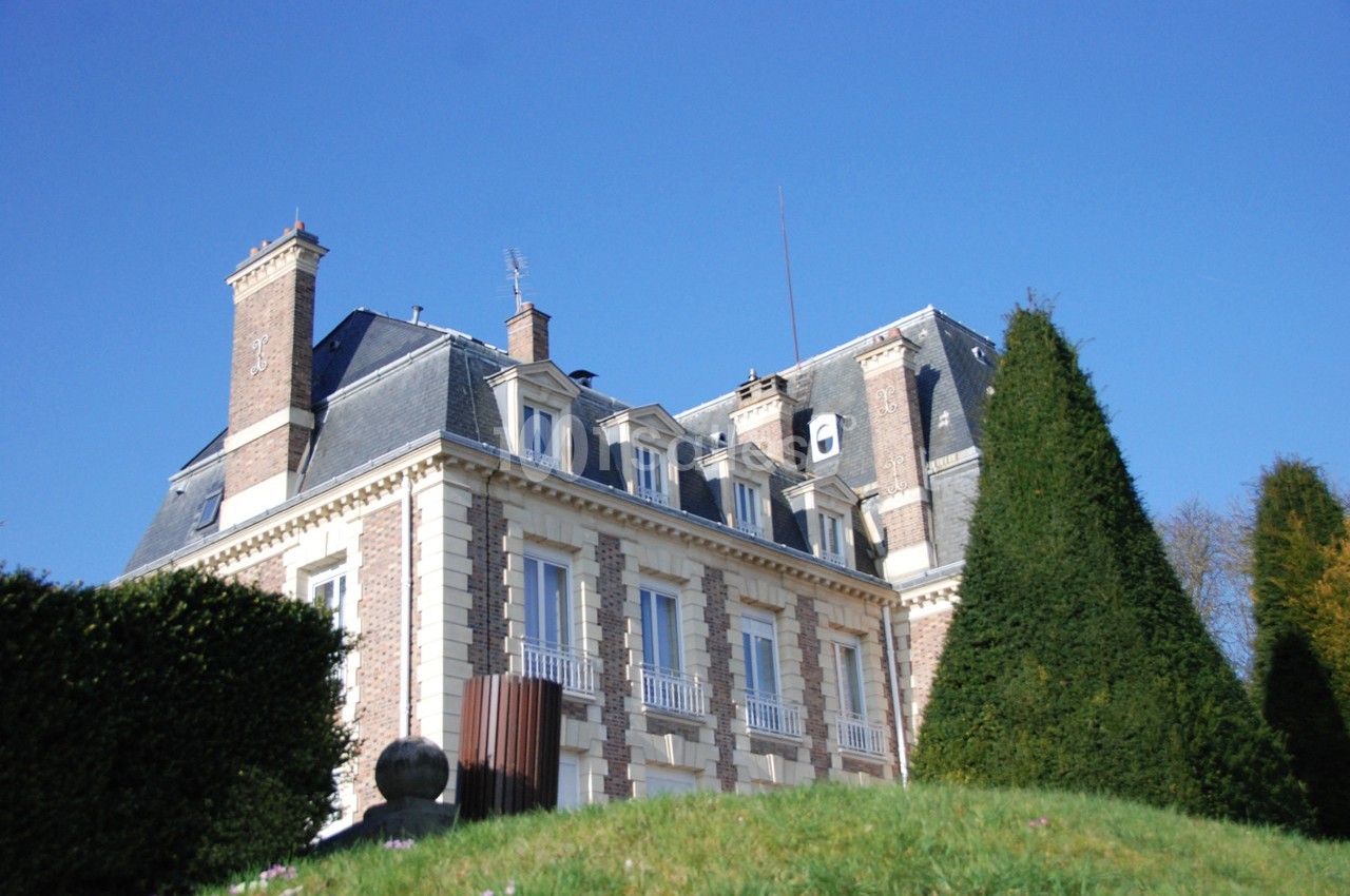 Façade d'un bâtiment en briques et pierres avec toit mansardé, entouré de haies et d'arbres sous un ciel bleu.