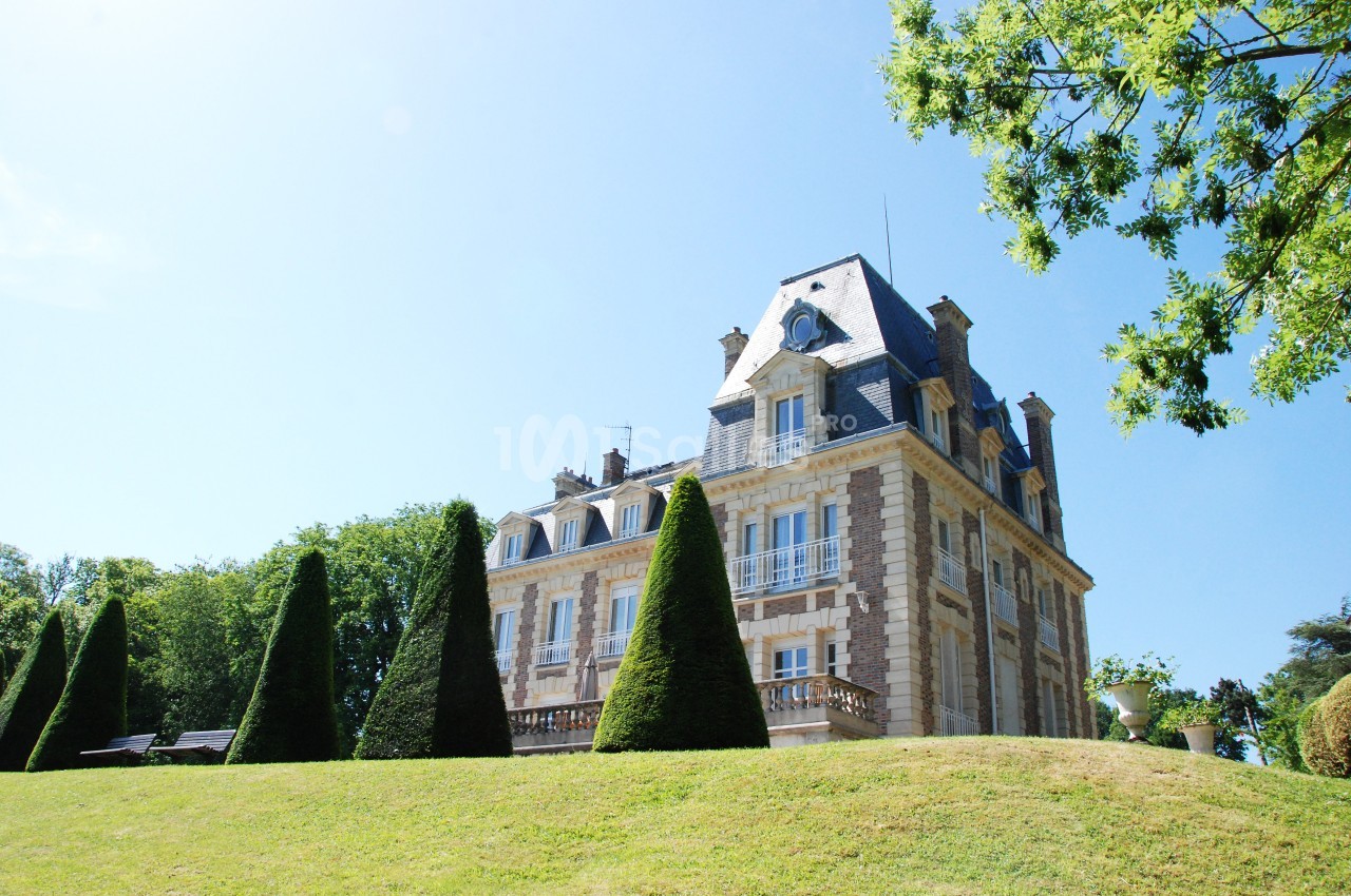 Manoir en pierre entouré d'arbres et de pelouses bien entretenues sous un ciel bleu clair.