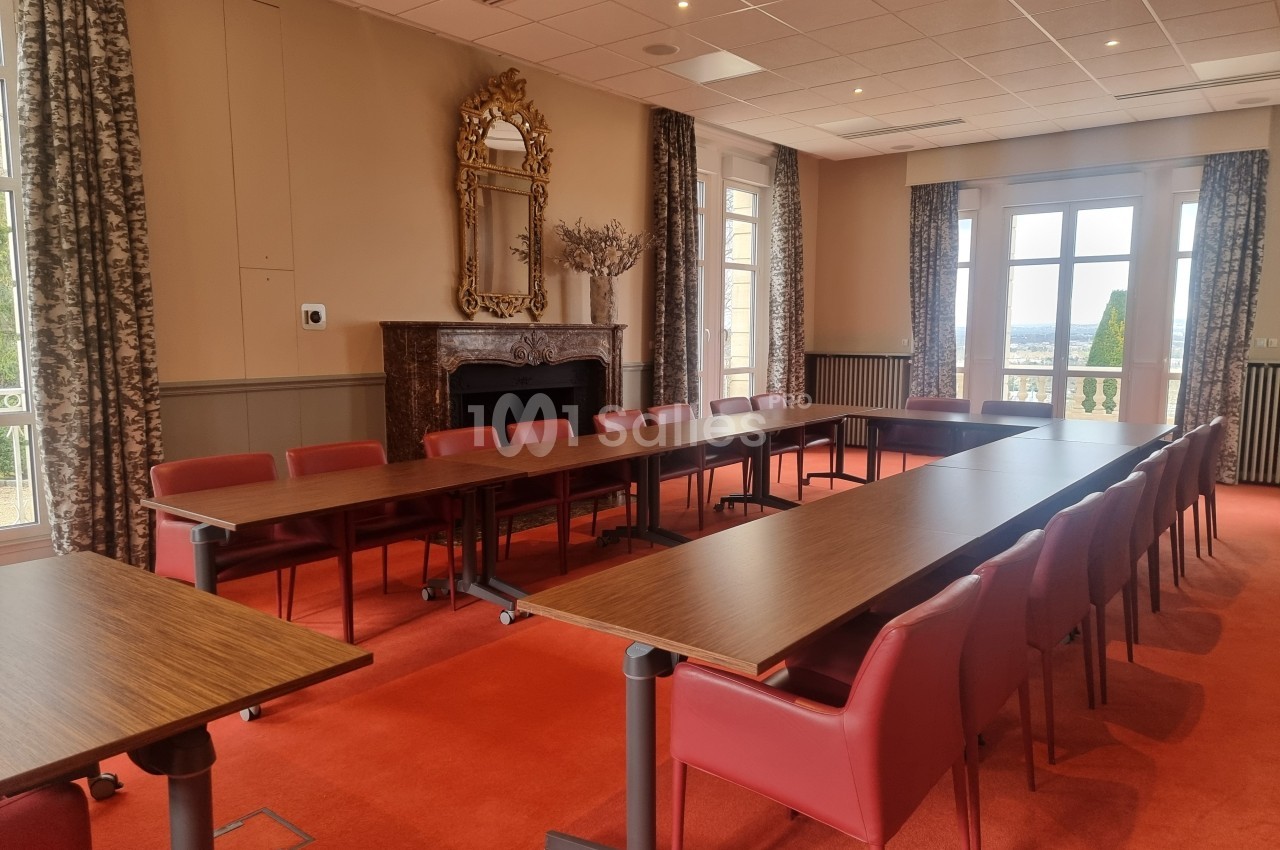 Salle de réunion lumineuse avec tables en bois, chaises rouges, cheminée, miroir et grandes fenêtres donnant sur l'extérieur.