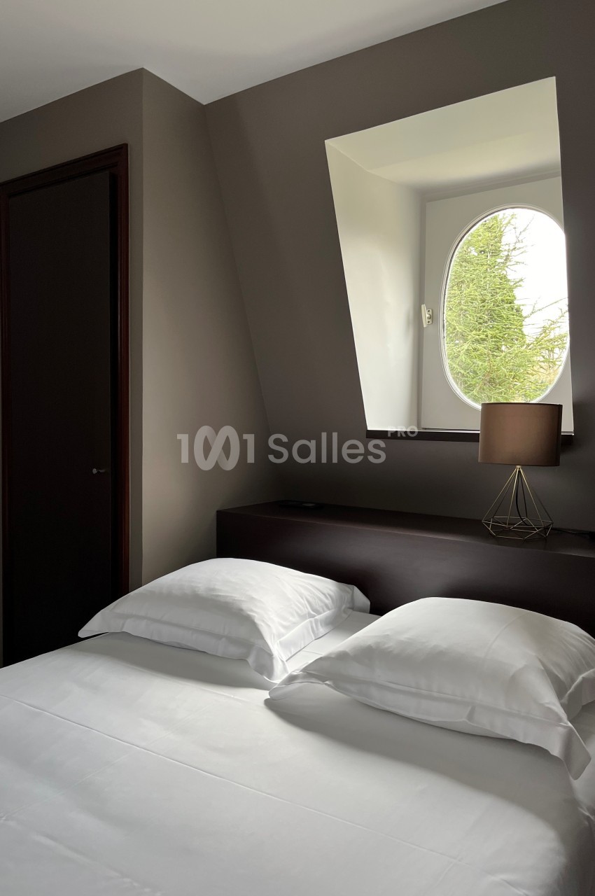 Lit double avec draps blancs dans une chambre mansardée, éclairée par une fenêtre ovale avec vue sur des arbres.