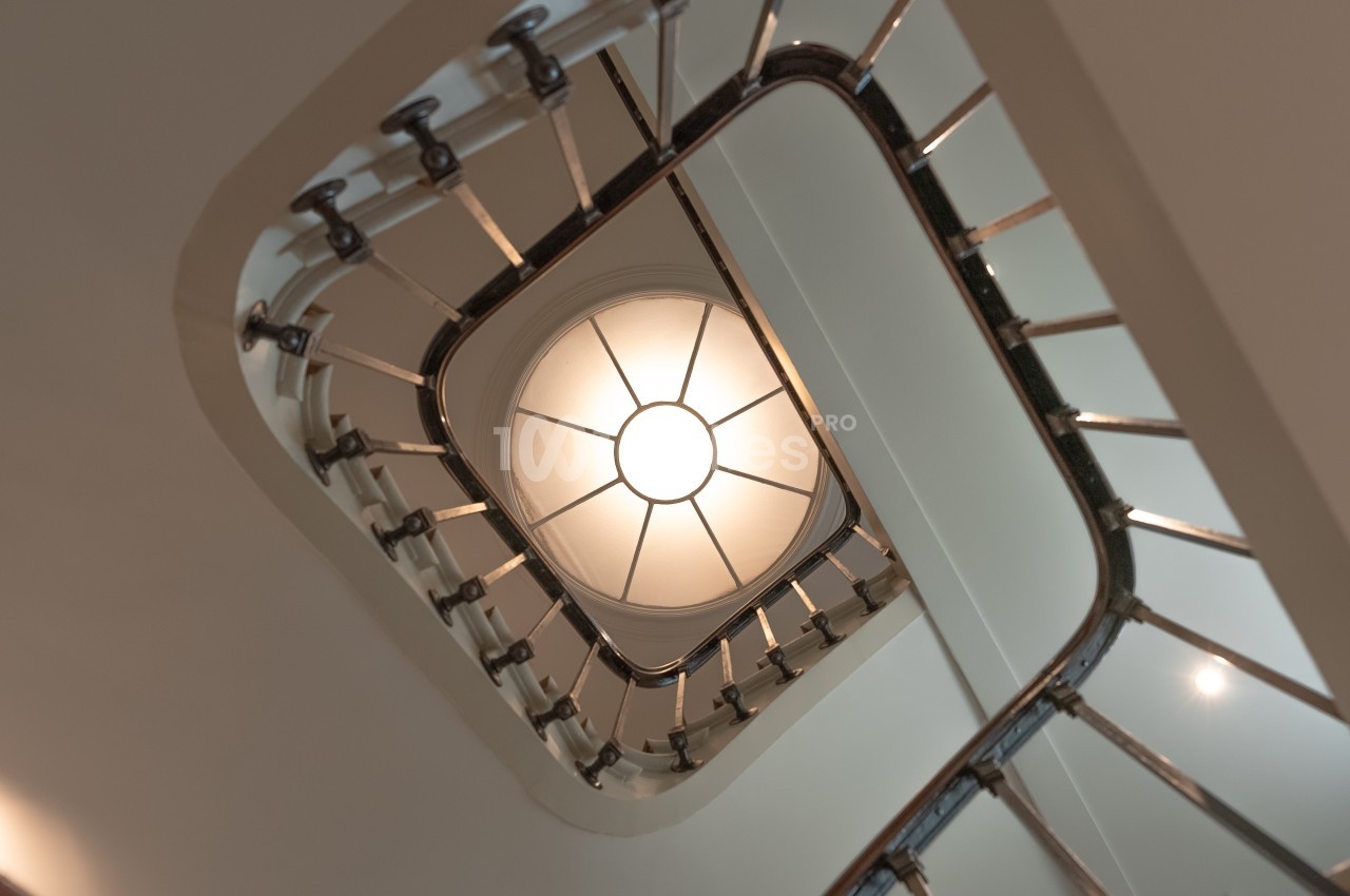 Vue en contre-plongée d'une cage d'escalier en colimaçon avec une rampe métallique et un plafond lumineux au centre.