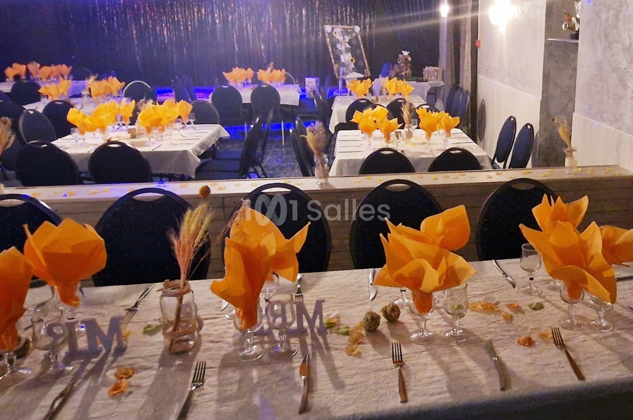 Salle de réception décorée avec des tables dressées, serviettes orange pliées et éléments décoratifs automnaux.
