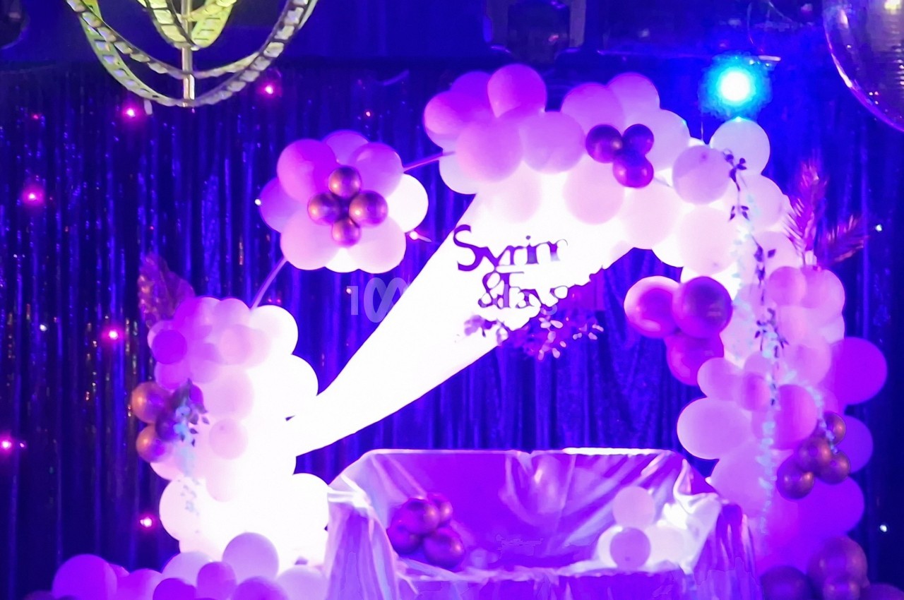 Décor festif avec arche de ballons roses et violets, éclairage coloré et rideau scintillant en arrière-plan.