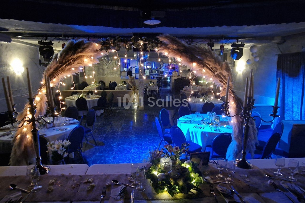 Salle de réception décorée avec des guirlandes lumineuses, des pampas et des tables dressées pour un événement.