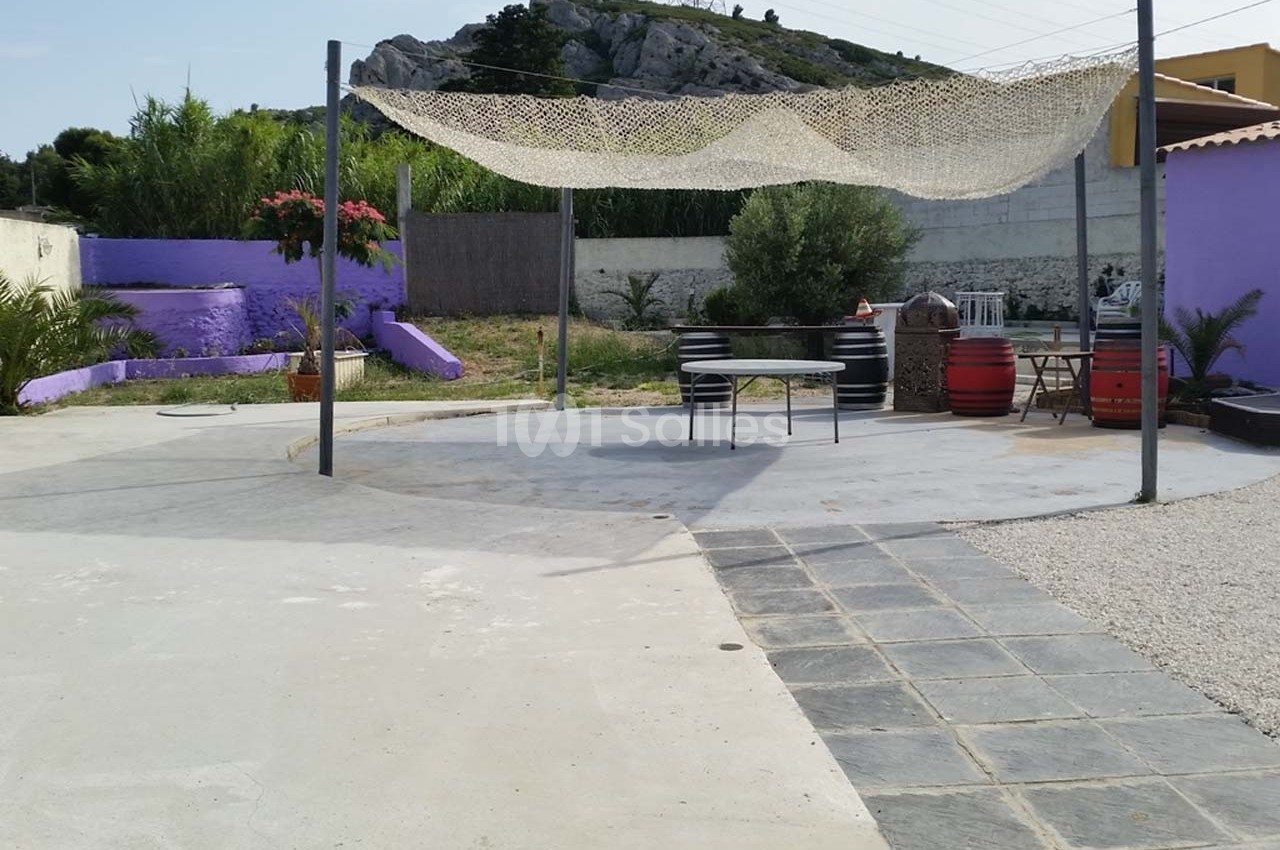 Terrasse extérieure avec voile d'ombrage, table, chaises, murs violets, végétation et colline en arrière-plan.