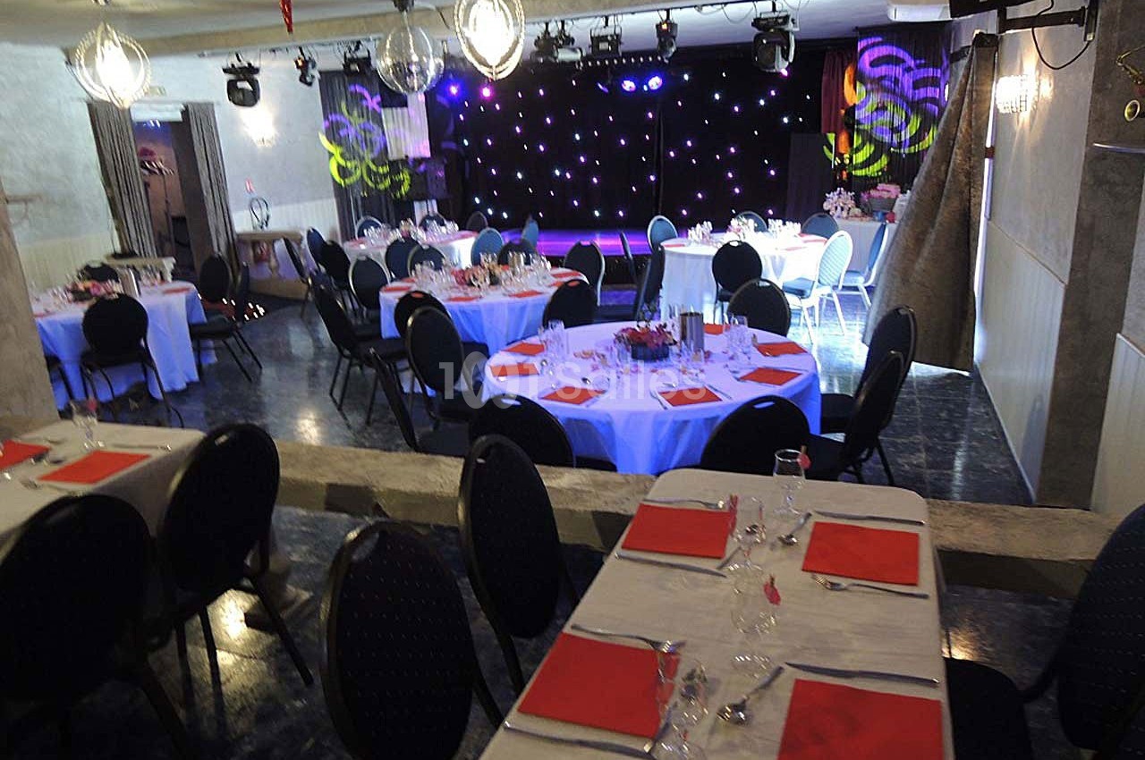 Salle de réception décorée avec des tables rondes et rectangulaires, nappes blanches et serviettes rouges, scène en arrière…