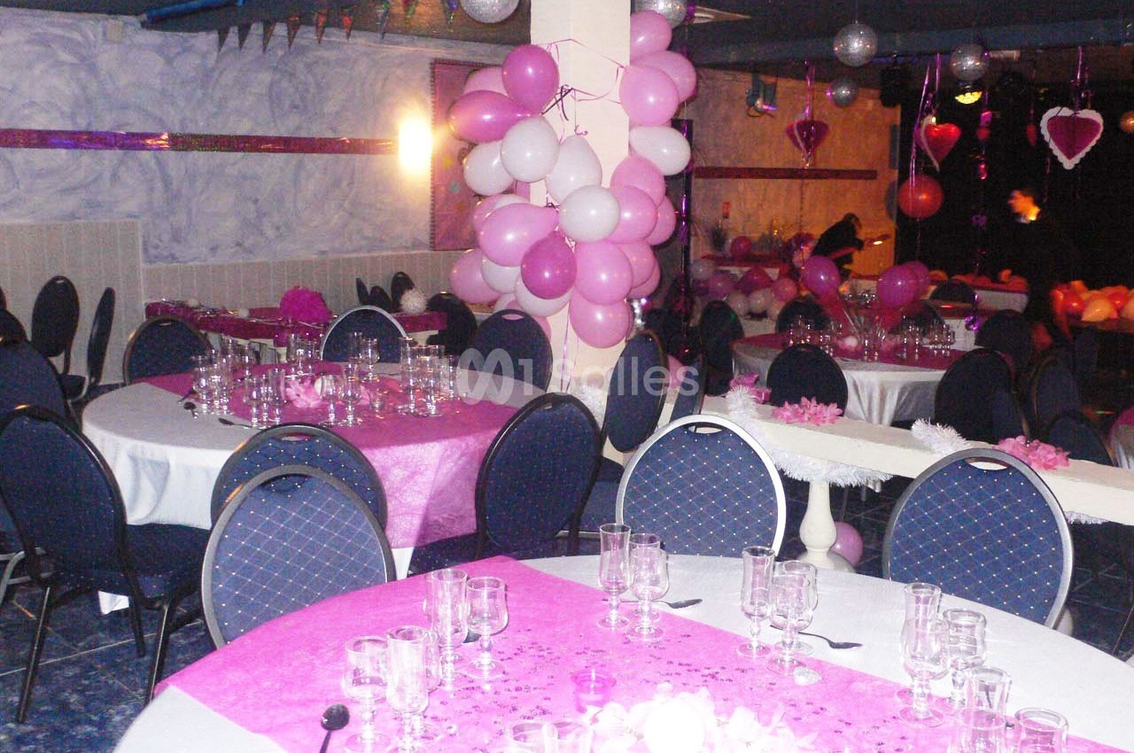 Salle décorée pour une fête avec des ballons roses et blancs, nappes roses, tables dressées et chaises bleues.