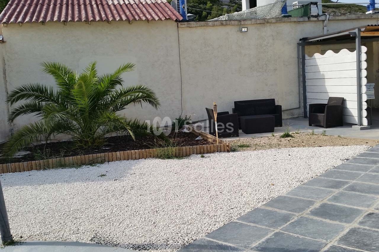 Cour extérieure avec gravier blanc, petit palmier, mobilier de jardin en rotin et mur beige en arrière-plan.