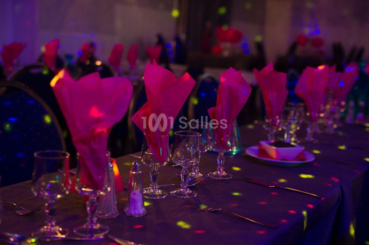Table dressée avec nappes sombres, serviettes roses pliées, verres et couverts, éclairée par des lumières colorées.