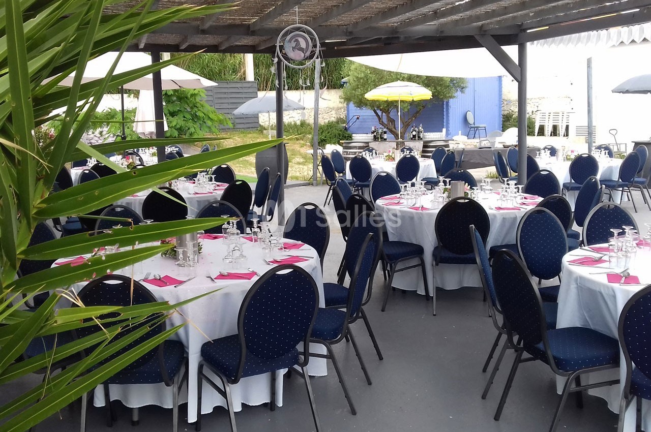 Tables rondes dressées avec nappes blanches et chaises bleues sous une pergola, dans un espace extérieur aménagé.