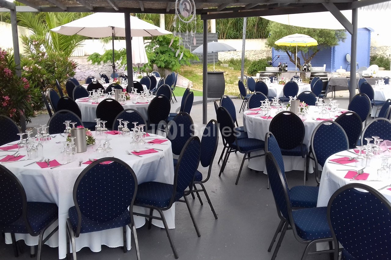 Salle extérieure aménagée avec des tables rondes dressées, nappes blanches, serviettes roses et chaises bleues.