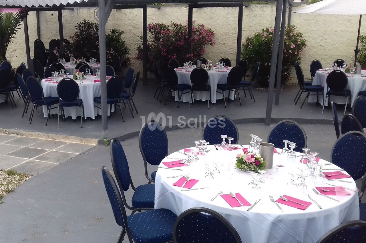 Tables rondes dressées avec nappes blanches, serviettes roses et verres, disposées dans un espace extérieur couvert.