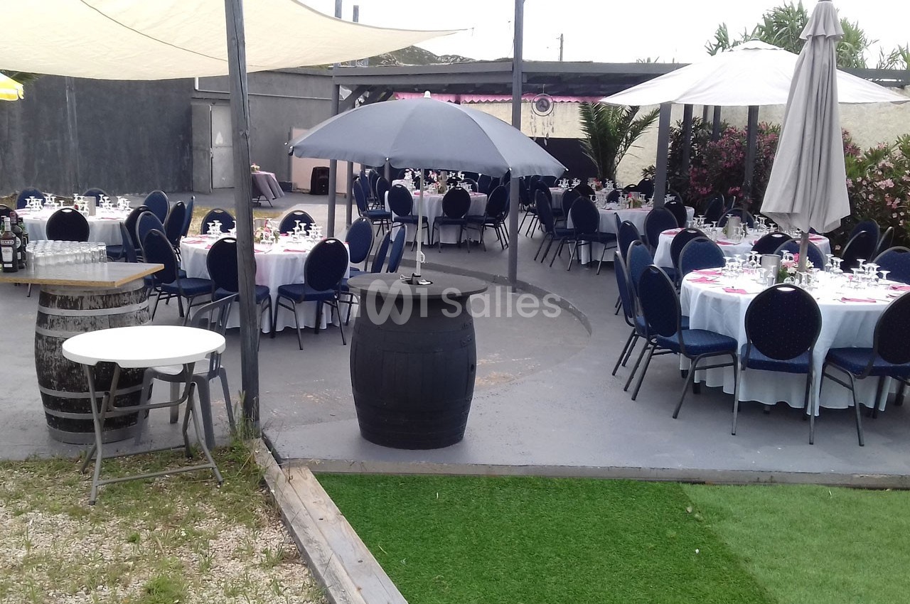 Terrasse aménagée avec des tables rondes dressées, des chaises bleues et des parasols, entourée de végétation.