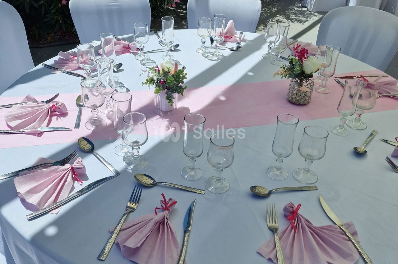 Table ronde dressée avec nappes blanches, serviettes pliées roses, couverts argentés et décorations florales au centre.