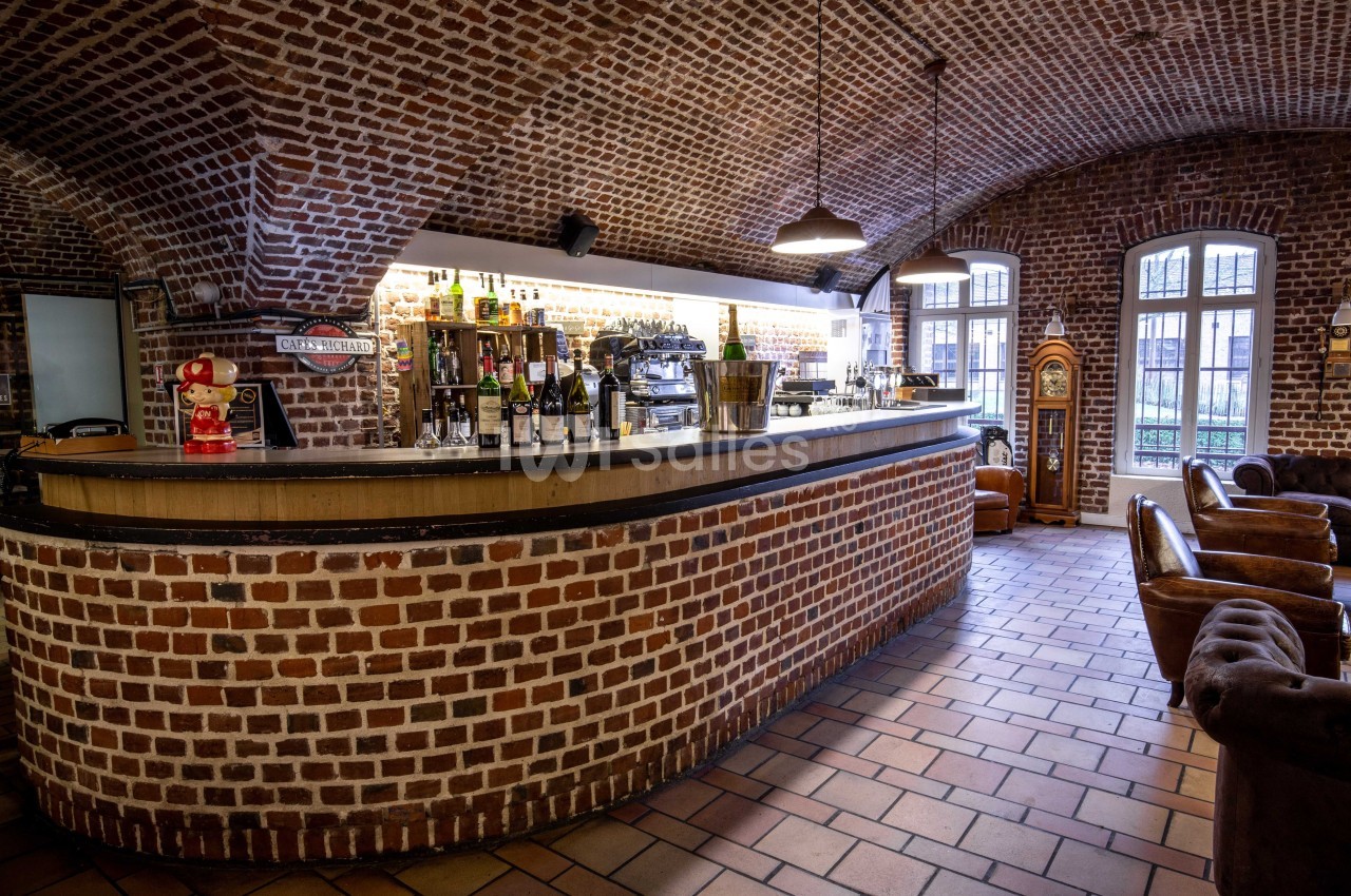 Bar en briques avec comptoir en bois, étagères de bouteilles, fauteuils en cuir et lumière naturelle par des fenêtres.