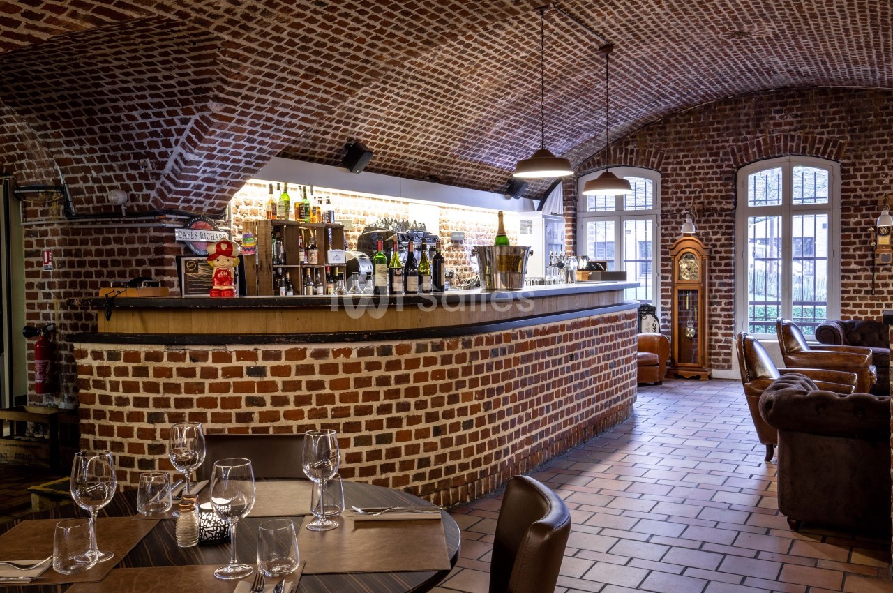 Bar en briques apparentes avec comptoir en bois, tables dressées au premier plan et fauteuils en cuir en arrière-plan.