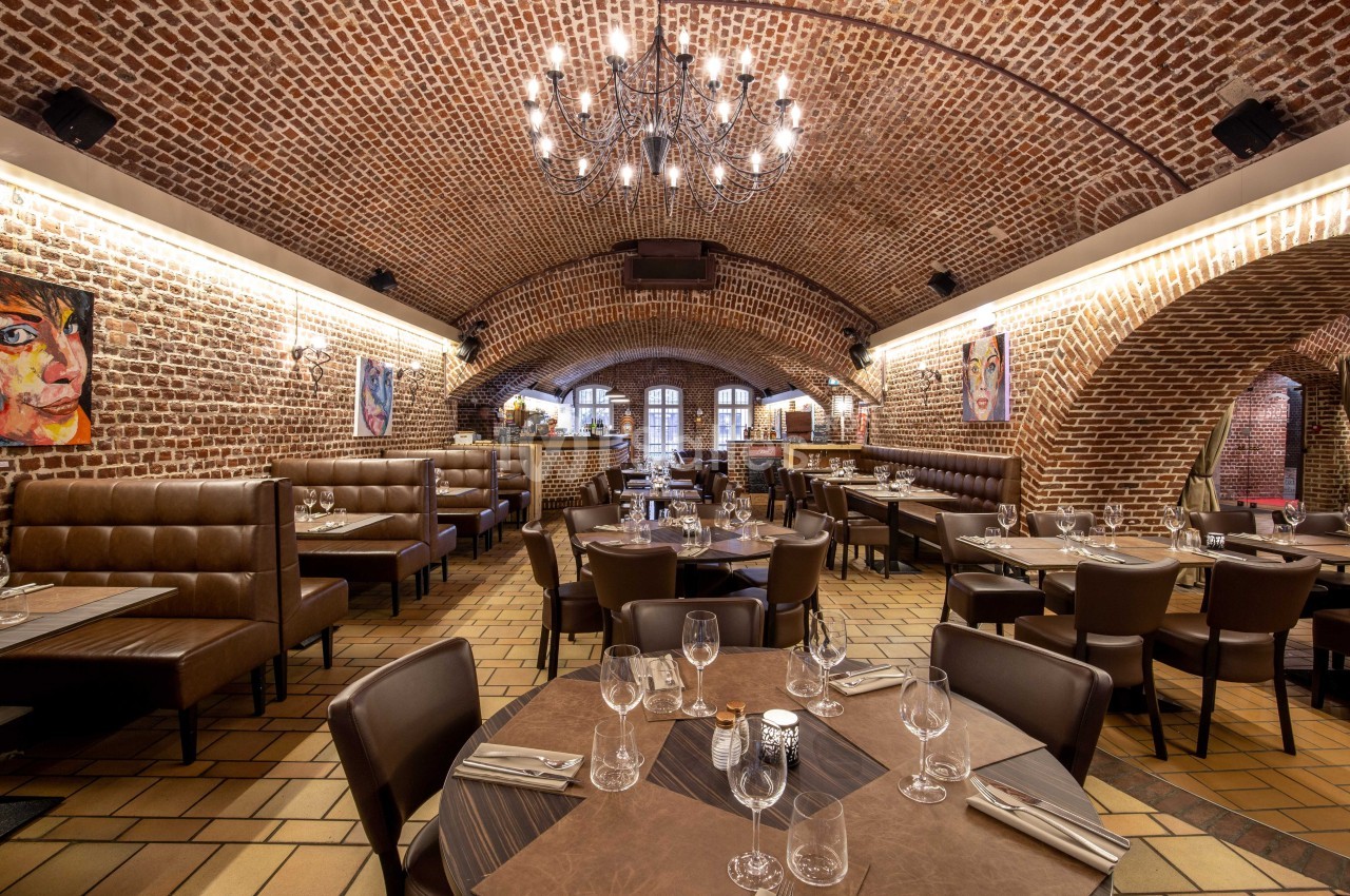 Salle de restaurant avec voûtes en briques, tables dressées, banquettes en cuir et éclairage chaleureux.