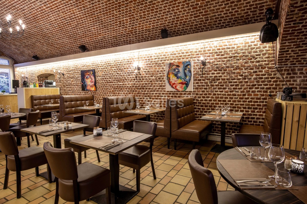 Salle de restaurant avec murs en briques, tables dressées, banquettes en cuir et tableaux colorés accrochés au mur.