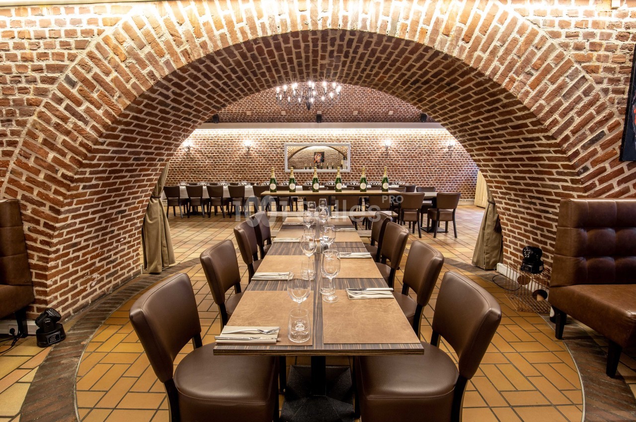 Salle de restaurant voûtée en briques avec tables dressées, chaises en cuir et éclairage chaleureux.