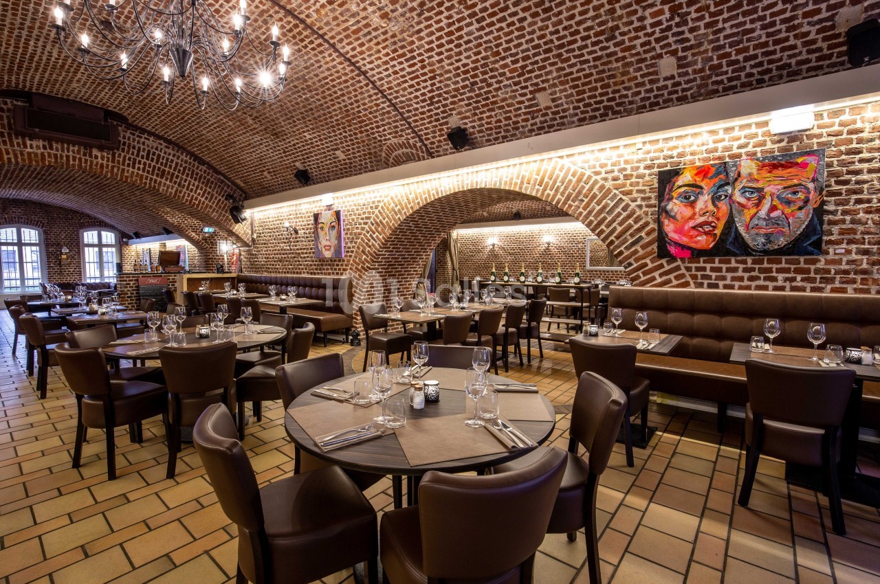 Salle de restaurant avec murs en briques, plafond voûté, tables dressées et tableaux colorés accrochés aux murs.