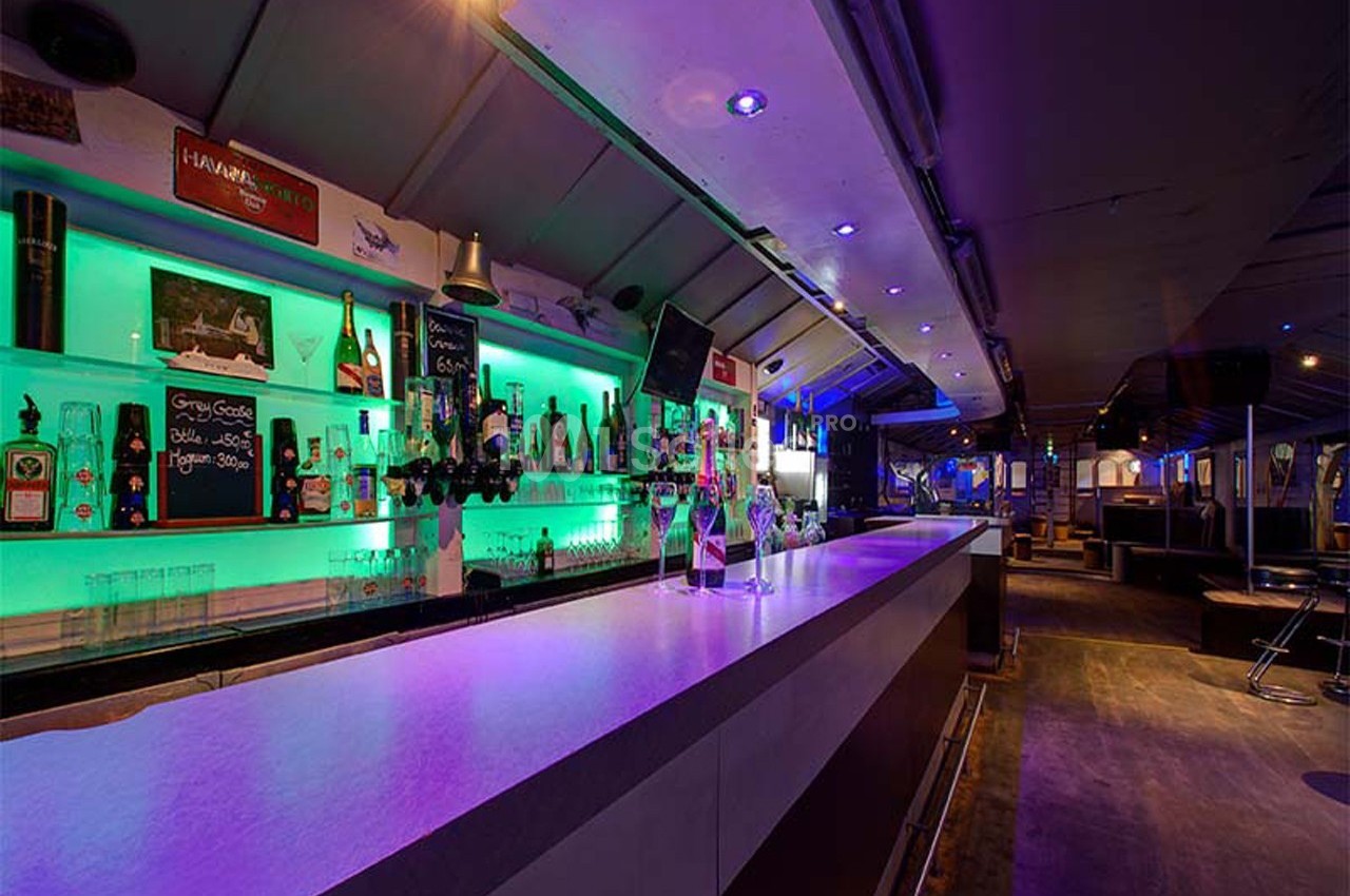 Bar moderne avec éclairage néon vert, comptoir long et rangées de bouteilles alignées sur des étagères.