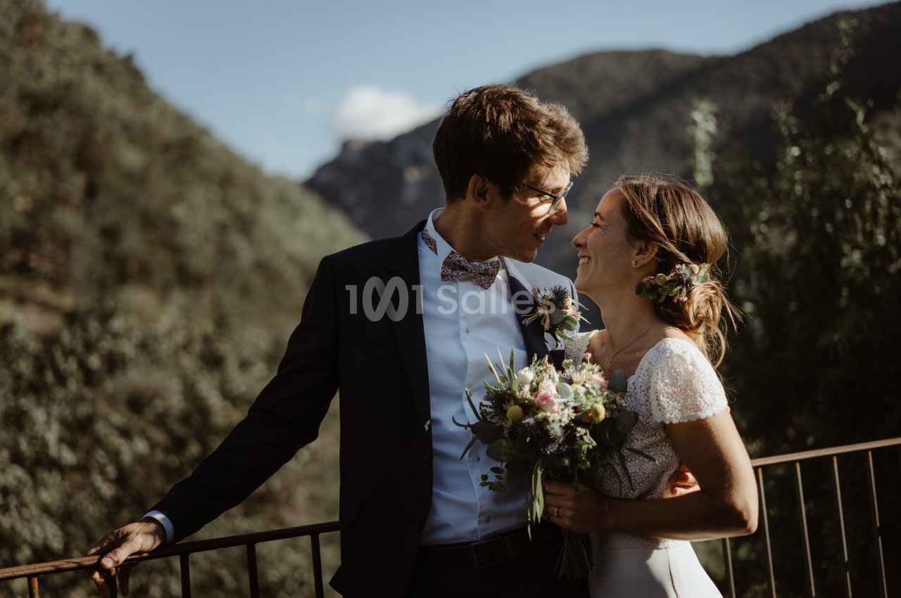 Un couple souriant en tenue de mariage se tient sur une terrasse avec un paysage montagneux en arrière-plan.
