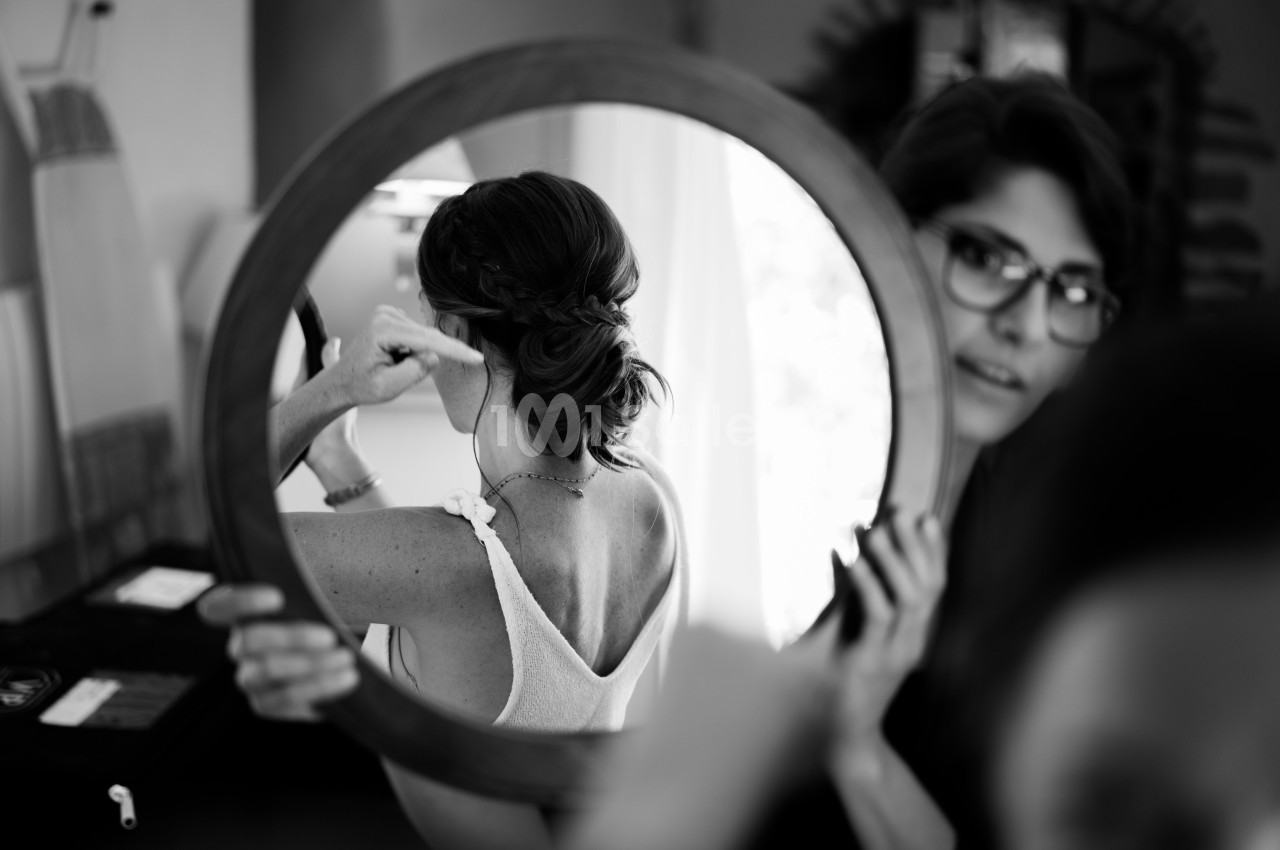Une femme ajuste sa coiffure devant un miroir rond, reflétant son dos et son chignon tressé.