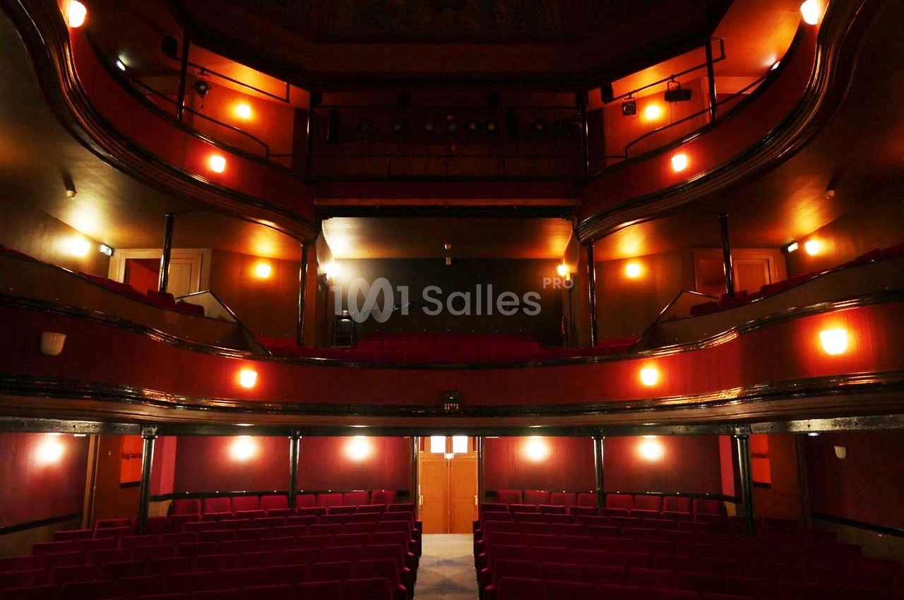Salle de théâtre vide avec sièges rouges, balcons en bois et éclairage tamisé.