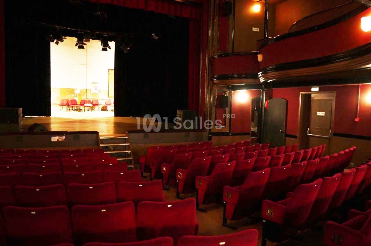 Salle de théâtre vide avec des rangées de fauteuils rouges, une scène éclairée et des murs sombres.