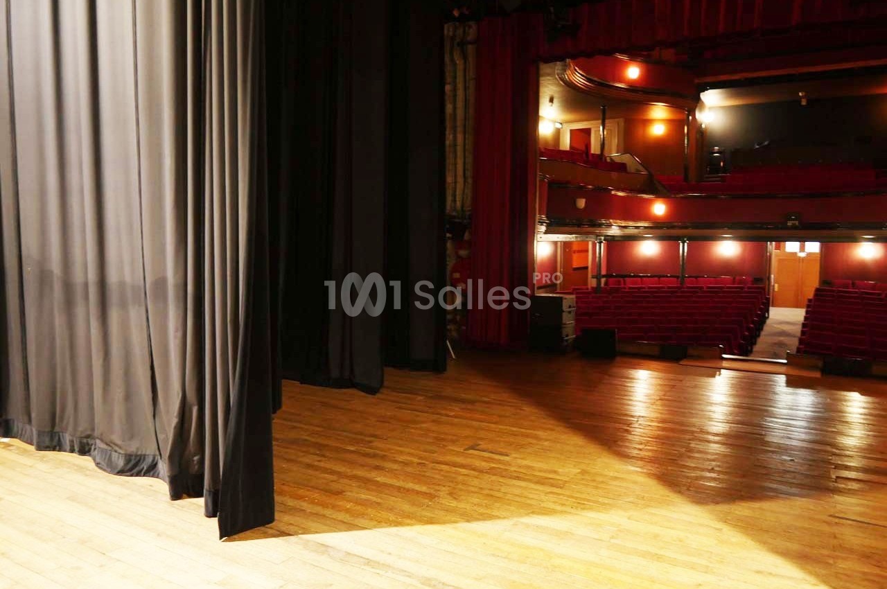 Vue depuis les coulisses d'une scène de théâtre avec rideaux noirs, plancher en bois et salle vide éclairée.