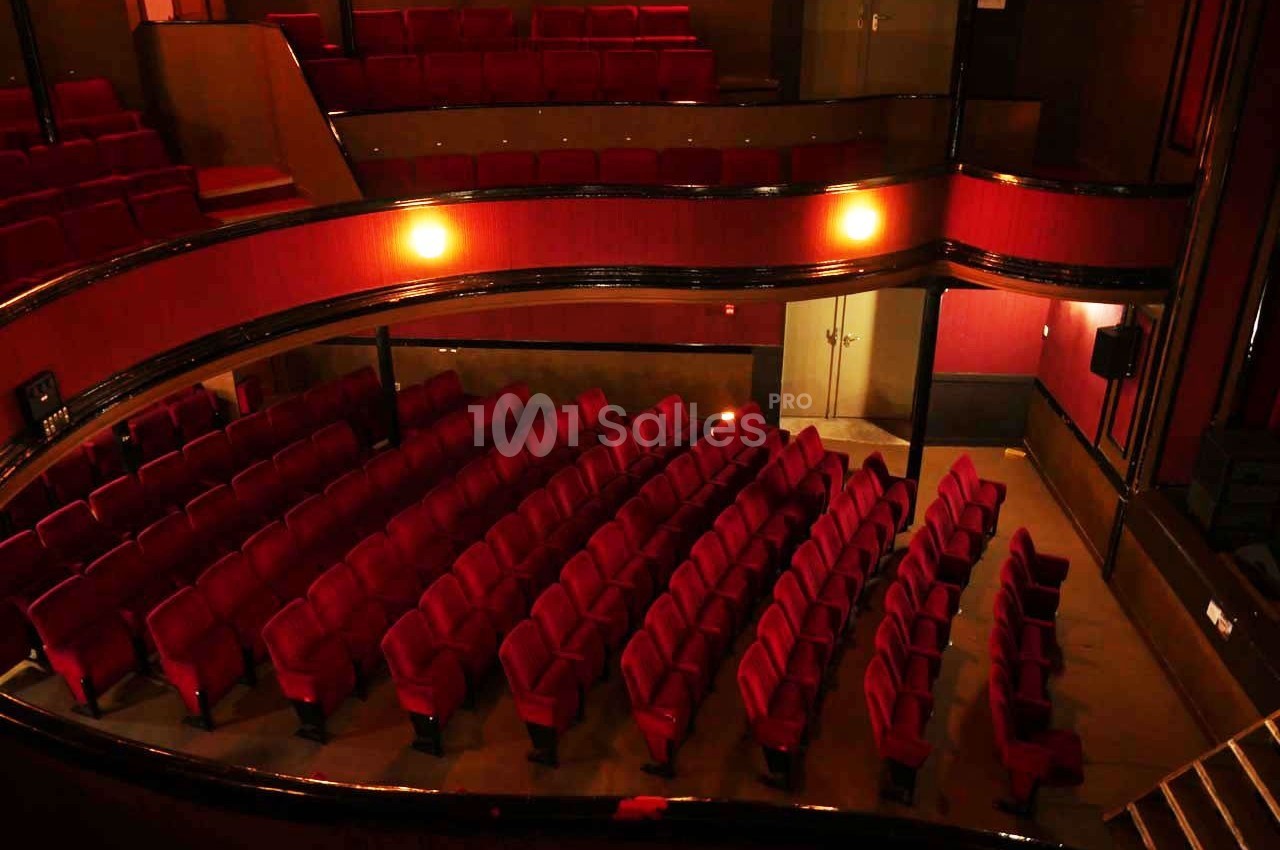 Salle de théâtre vide avec des rangées de sièges rouges et un éclairage tamisé.