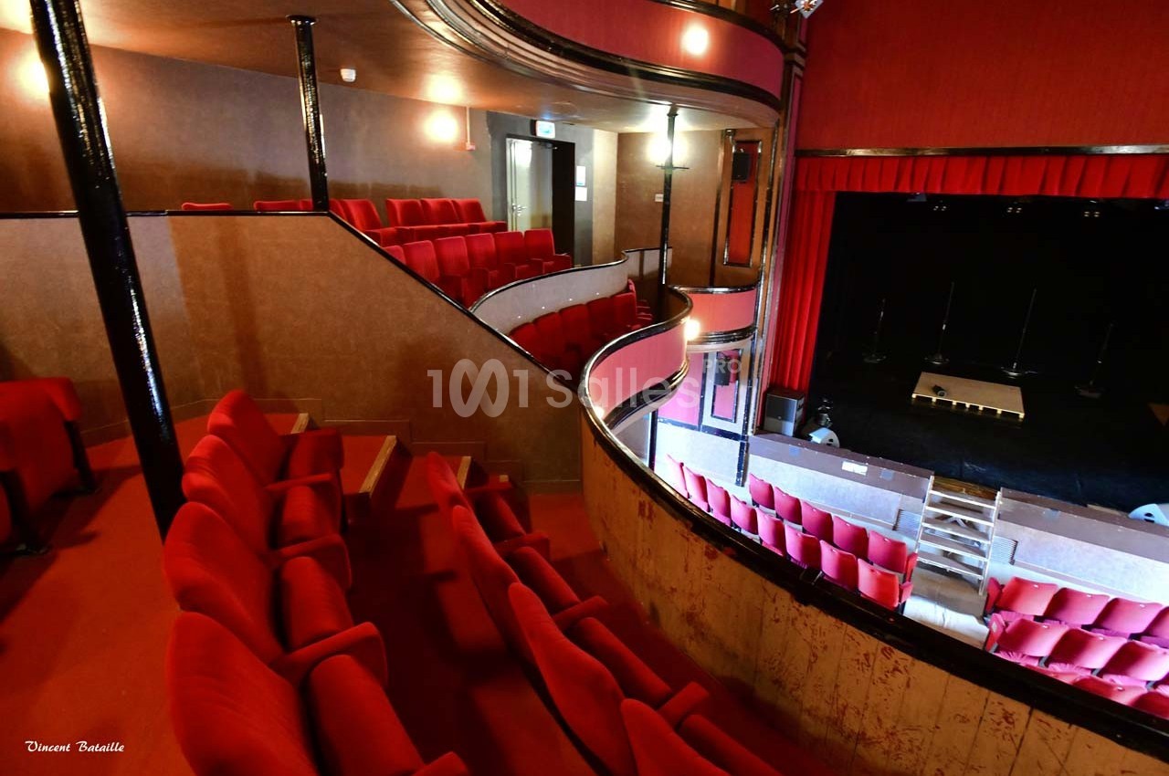 Salle de théâtre avec sièges rouges, balcons en bois et scène vide éclairée par une lumière tamisée.