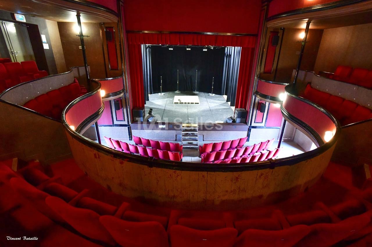 Salle de théâtre vide avec sièges rouges, scène éclairée et rideaux noirs, vue depuis le balcon.