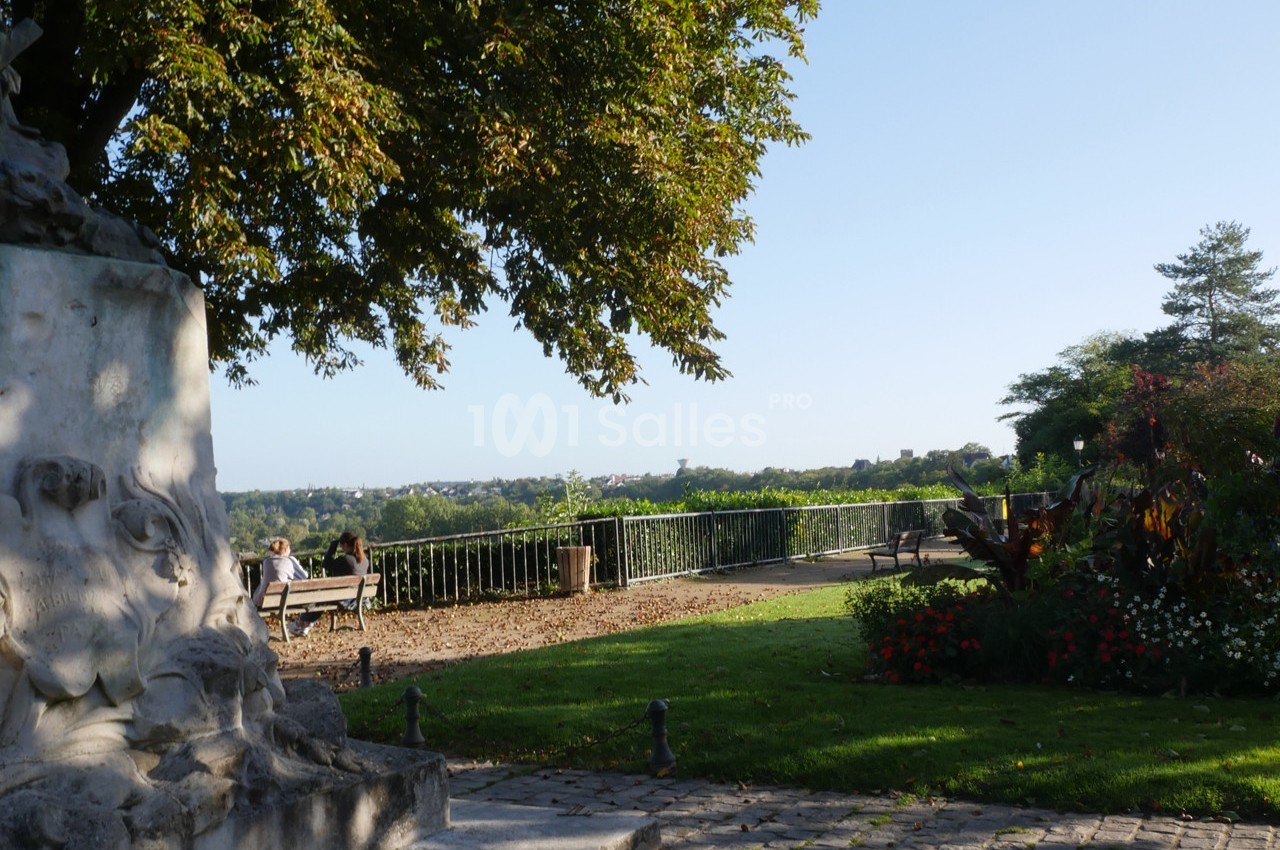Vue d'un parc avec des bancs, des fleurs, une statue partielle et un panorama sur un paysage verdoyant à l'horizon.
