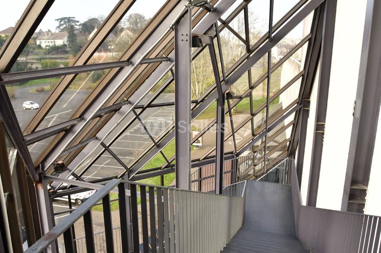 Escalier métallique intérieur avec vue sur un parking et des espaces verts à travers une structure vitrée inclinée.