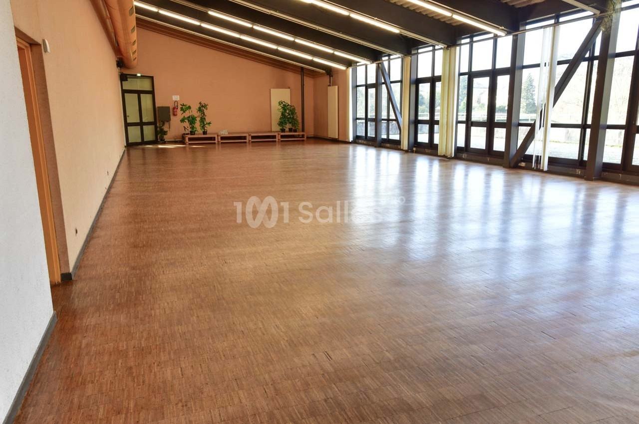 Salle spacieuse avec parquet en bois, grandes baies vitrées et quelques plantes en arrière-plan.