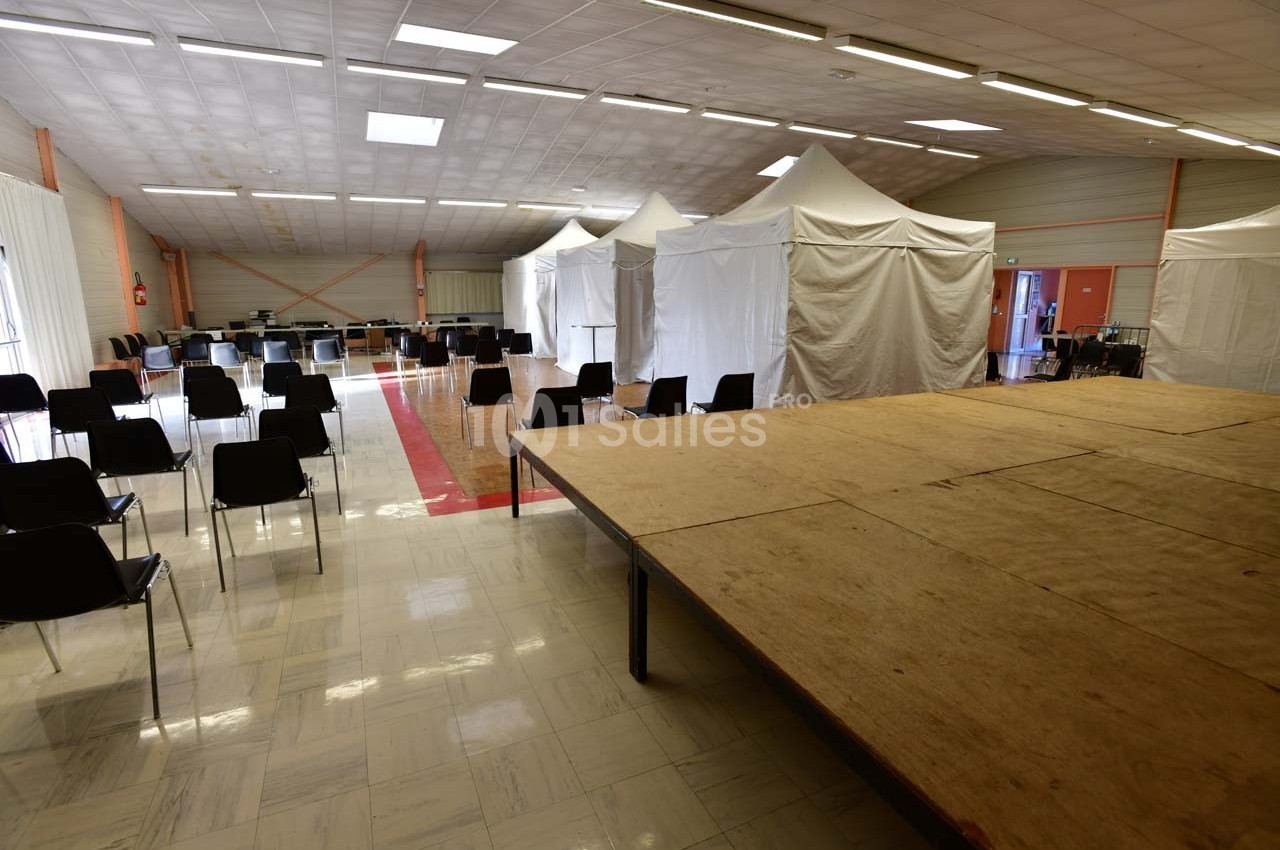 Salle avec une estrade en bois, des chaises disposées en rangées et des tentes blanches alignées en arrière-plan.