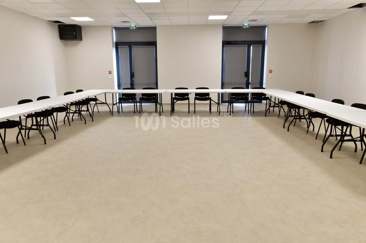 Salle de réunion vide avec des tables blanches disposées en U et des chaises noires, éclairée par des lumières au plafond.