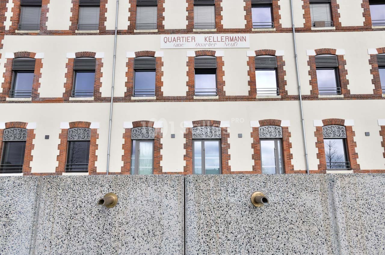 Façade d'un bâtiment en briques et crépi avec des fenêtres alignées, vue derrière un mur en béton.