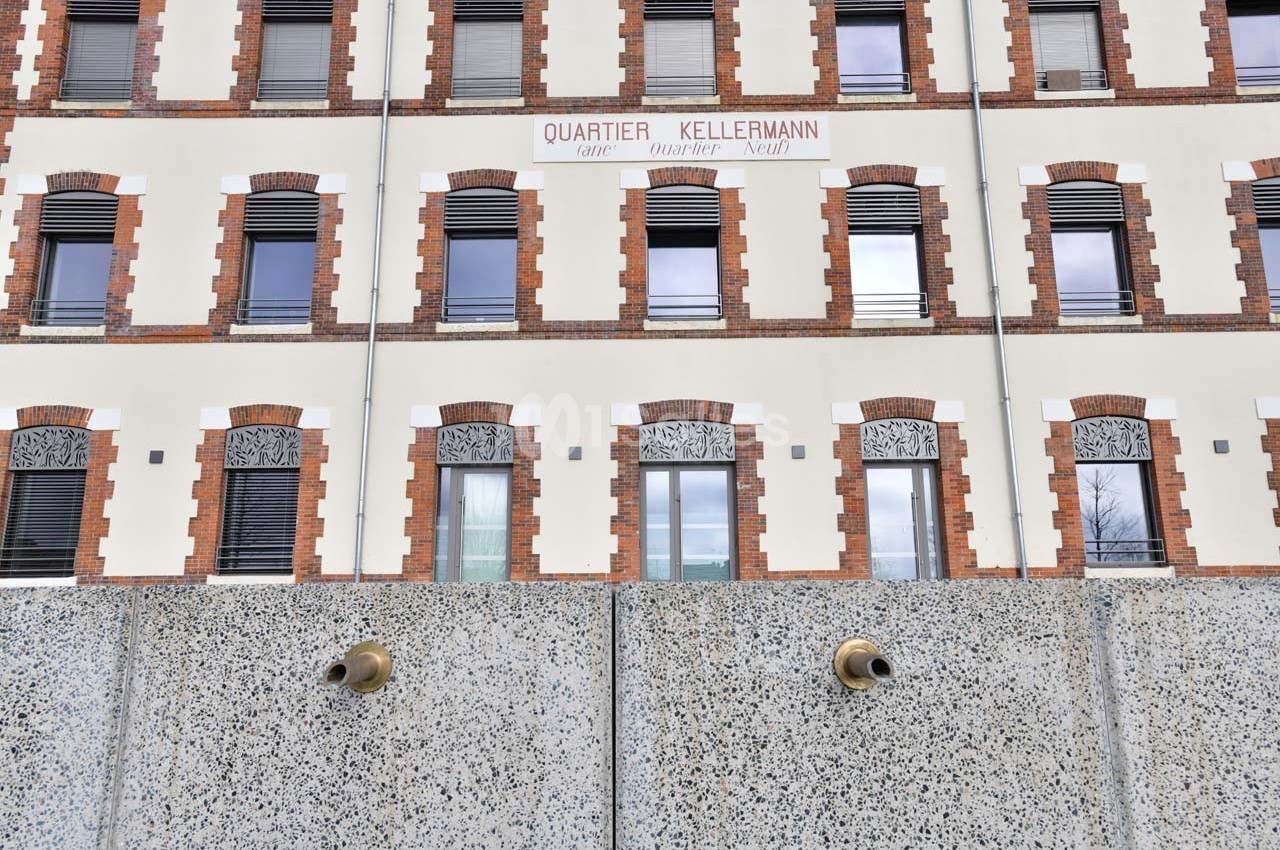 Façade d'un bâtiment en briques et crépi avec des fenêtres alignées, vue derrière un mur en béton percé de tuyaux.