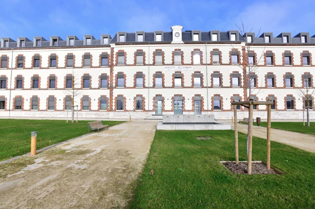 Façade d'un bâtiment rénové en briques blanches et rouges, entouré d'une pelouse et de chemins piétonniers.